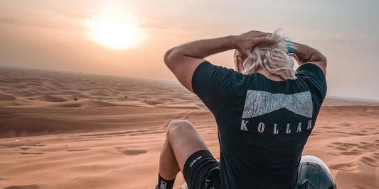 A blonde-haired man wearing a black Kollar t-shirt sits on the sand in a vast desert, gazing at the sunrise with his hands on his head. A motorcycle helmet rests beside him, symbolizing a 