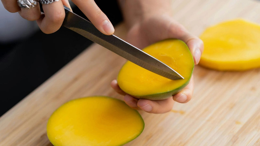 Person holding a mango in their palm while slicing it with a knife to prepare a recipe.